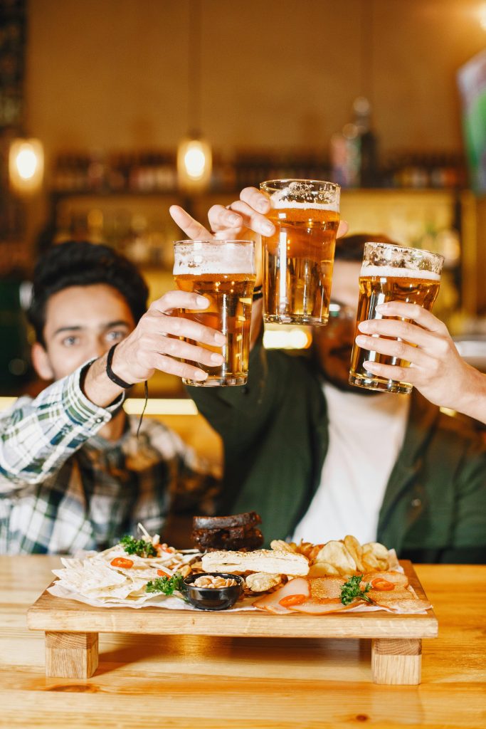 Three friends clinking their glass of beers with a table of food in the front.
